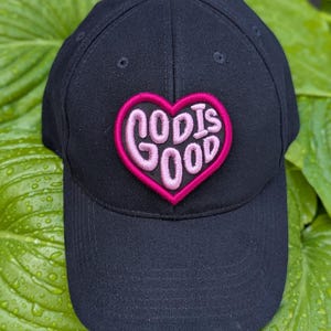 May include: Black baseball cap with a pink heart-shaped patch. The patch features the words "GOD IS GOOD" in a stylized, cursive font. The cap is set against a backdrop of green leaves.