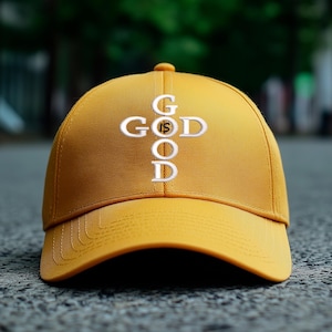 Puede incluir: Gorra de béisbol de color amarillo mostaza con la inscripción "GOD IS GOOD" bordada en blanco. La gorra tiene una visera curva y un botón en la parte superior. El texto está dispuesto en forma de cruz.
