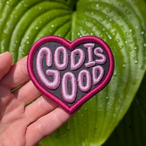May include: A heart-shaped patch with the words "GOD IS GOOD" embroidered in pink thread on a black background. The patch has a dark pink border. The background is a blurred image of green leaves.