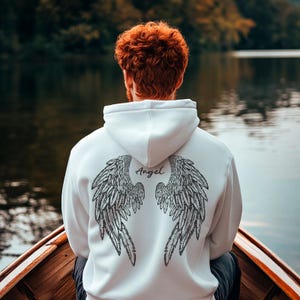 Puede incluir: Sudadera con capucha blanca con un diseño de alas de ángel negras en la espalda. La palabra "Angel" está escrita encima de las alas. La persona está sentada en un bote de madera en un lago.