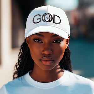 Può includere: Cappellino da baseball bianco con la scritta "GOD IS" in nero. Il cappellino è indossato da una persona che indossa una maglietta bianca. L'immagine è un primo piano.