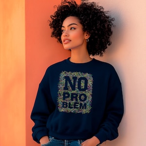 Può includere: Una felpa blu navy con la scritta "NO PROBLEM" in un design colorato e scarabocchiato. La felpa è indossata da una persona con i capelli ricci scuri, in piedi davanti a un muro color pesca e bianco.