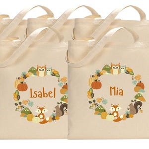 May include: Set of six beige canvas tote bags with brown handles. Each bag features a wreath design with owls, squirrels, and pumpkins. The bags are personalized with the names "Isabel" and "Mia".