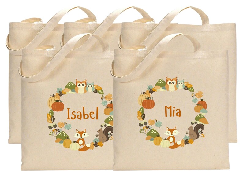 May include: Set of six beige canvas tote bags with brown handles. Each bag features a wreath design with owls, squirrels, and pumpkins. The bags are personalized with the names "Isabel" and "Mia".