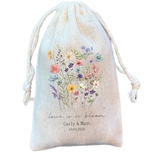 May include: A beige drawstring bag with a floral design. The bag features a bouquet of colourful wildflowers in pink, blue, yellow, and purple. Text on the bag reads "love is in bloom", with names and a date below.