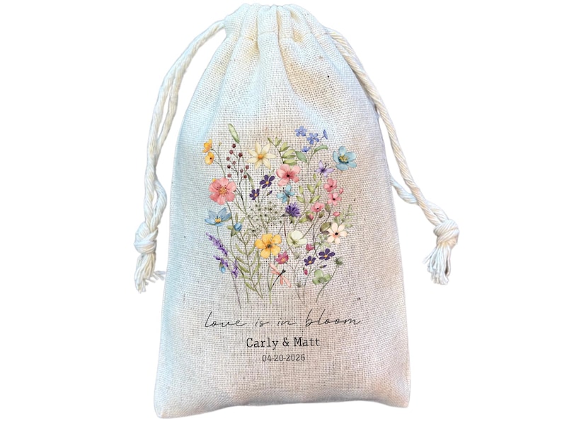 May include: A beige drawstring bag with a floral design. The bag features a bouquet of colourful wildflowers in pink, blue, yellow, and purple. Text on the bag reads "love is in bloom", with names and a date below.