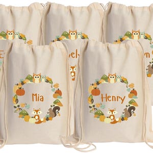 May include: Five white canvas drawstring bags with a woodland animal theme. Each bag has a different name printed on it: Mia, Henry, Evelyn, Owen, and Ethan. The bags feature a circle of woodland animals including owls, foxes, squirrels, and mushrooms.