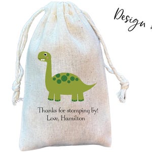 May include: A beige drawstring pouch made of woven fabric featuring a green dinosaur illustration with dark green spots. Below the dinosaur, the text reads: Thanks for stomping by! Love, Hamilton. The top right corner displays the text Design #5.