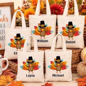 May include: Five off-white canvas tote bags, each featuring a cartoon turkey wearing a black pilgrim hat and colorful tail feathers. Each bag is personalized with a name printed in black text below the turkey graphic.