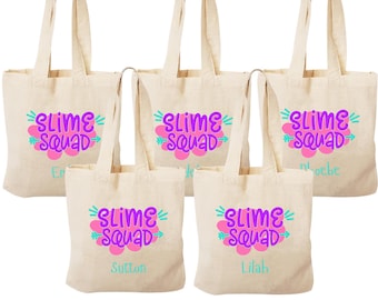 May include: Five white canvas tote bags with purple and pink "Slime Squad" text and a turquoise arrow. Each bag has a different name on it: Emma, Sutton, Lilah, Phoebe, and another name that is unclear.