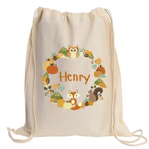 May include: A beige drawstring bag with a personalized design featuring a wreath of autumn leaves, pumpkins, owls, a fox, and a squirrel. The name "Henry" is printed in orange in the center of the wreath.