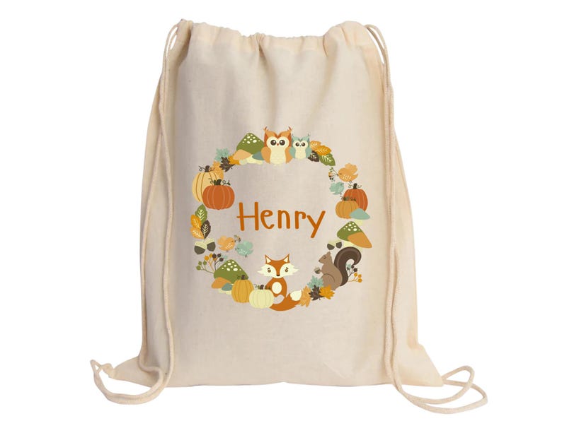 May include: A beige drawstring bag with a personalized design featuring a wreath of autumn leaves, pumpkins, owls, a fox, and a squirrel. The name "Henry" is printed in orange in the center of the wreath.