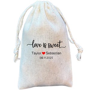 May include: A white cotton drawstring bag with the text "love is sweet" printed in black. Below the text are the names "Taylor" and "Sebastian" with a red heart symbol between them. The date "06.11.2025" is printed below the names.