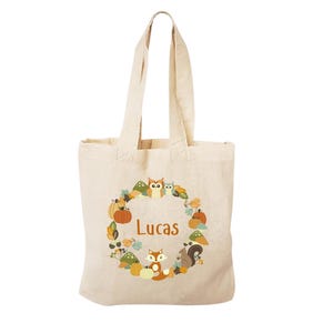 May include: A beige canvas tote bag with a brown, orange, and yellow autumn-themed design. The design features a wreath of leaves, pumpkins, owls, squirrels, and a fox. The name "Lucas" is printed in orange in the center of the wreath.