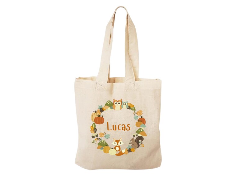 May include: A beige canvas tote bag with a brown, orange, and yellow autumn-themed design. The design features a wreath of leaves, pumpkins, owls, squirrels, and a fox. The name "Lucas" is printed in orange in the center of the wreath.
