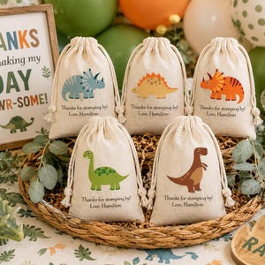 May include: Five beige drawstring favor bags featuring colorful dinosaur illustrations and the printed text Thanks for stomping by! Love, Hamilton. The bags are arranged in a woven basket, each displaying a different dinosaur species in various colors.