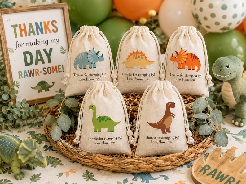 May include: Five beige drawstring favor bags featuring colorful dinosaur illustrations and the printed text Thanks for stomping by! Love, Hamilton. The bags are arranged in a woven basket, each displaying a different dinosaur species in various colors.