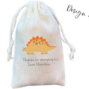 May include: A beige drawstring pouch made of woven fabric featuring a graphic of a tan dinosaur with orange plates on its back. Below the dinosaur, the text reads: Thanks for stomping by! Love, Hamilton. The upper right corner displays Design #3.