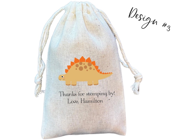 May include: A beige drawstring pouch made of woven fabric featuring a graphic of a tan dinosaur with orange plates on its back. Below the dinosaur, the text reads: Thanks for stomping by! Love, Hamilton. The upper right corner displays Design #3.