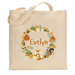 May include: A beige canvas tote bag with a fall-themed design featuring a fox, owls, pumpkins, and leaves. The bag has the name "Evelyn" printed in orange on the front.