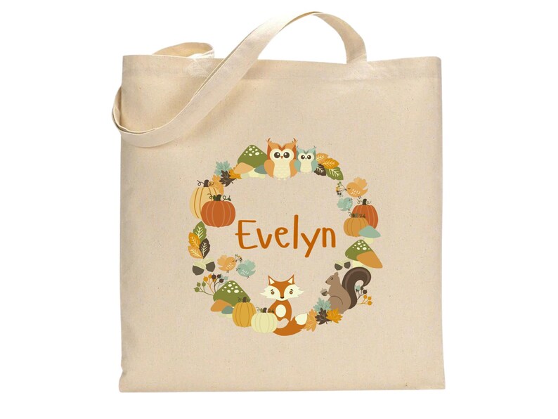 May include: A beige canvas tote bag with a fall-themed design featuring a fox, owls, pumpkins, and leaves. The bag has the name "Evelyn" printed in orange on the front.