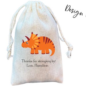 May include: A beige drawstring pouch made of woven fabric featuring a graphic of an orange triceratops dinosaur. Below the dinosaur, the text reads: Thanks for stomping by! Love, Hamilton. The top right corner displays the text Design #1.