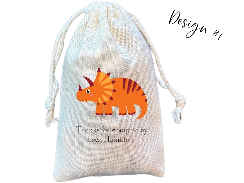 May include: A beige drawstring pouch made of woven fabric featuring a graphic of an orange triceratops dinosaur. Below the dinosaur, the text reads: Thanks for stomping by! Love, Hamilton. The top right corner displays the text Design #1.