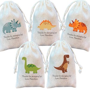 May include: Five off-white drawstring pouches made of fabric, each featuring a colorful cartoon dinosaur illustration and the printed text: Thanks for stomping by! Love, Hamilton. The dinosaurs include a blue triceratops, orange stegosaurus, orange triceratops, green long-neck, and brown t-rex.