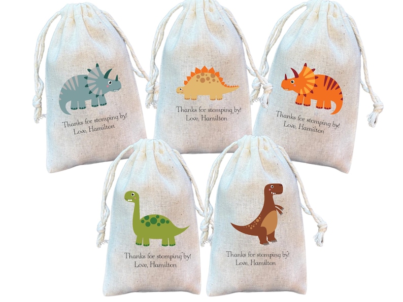 May include: Five off-white drawstring pouches made of fabric, each featuring a colorful cartoon dinosaur illustration and the printed text: Thanks for stomping by! Love, Hamilton. The dinosaurs include a blue triceratops, orange stegosaurus, orange triceratops, green long-neck, and brown t-rex.