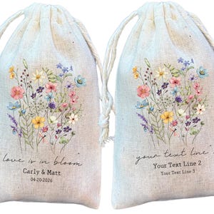 May include: Two beige drawstring favour bags featuring a watercolour floral design. Each bag has a bouquet of pink, blue, yellow, and purple flowers. One bag has the text "love is in bloom" with a name and date. The other has space for custom text.