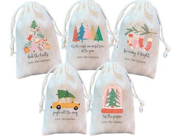 May include: Five beige drawstring gift bags with Christmas-themed designs. Each bag features a different festive illustration, including Christmas trees, ornaments, and a car carrying a tree. Text on the bags includes holiday phrases like "Deck the halls" and "Jingle all the way."