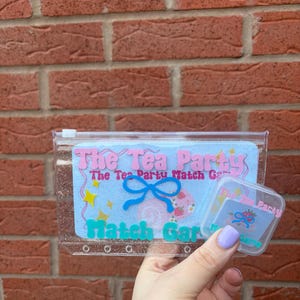 May include: A clear, zipped pouch containing a light blue card with the text "The Tea Party Match Game". The card features a blue bow and strawberry illustrations. A smaller, clear container with matching graphics is also present.
