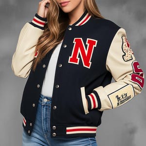 Puede incluir: Chaqueta universitaria azul marino y crema con una gran "N" roja en el pecho. La chaqueta presenta mangas color crema, puños y cuello a rayas rojas y blancas, y un parche en la manga. La chaqueta tiene cierre de botones a presión.