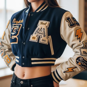 Puede incluir: Chaqueta universitaria azul marino y crema con múltiples parches bordados. La chaqueta presenta un gran parche con la letra "A", rayos y el texto "Garage 93". Las mangas son de color crema y la chaqueta tiene un corte corto.
