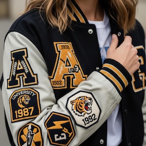 Op de afbeelding: Zwarte varsity jas met crèmekleurige mouwen en meerdere geborduurde patches. De jas heeft goud en zwart gestreepte manchetten en kraag. Patches zijn onder meer de letter "A" en een tijgerafbeelding.
