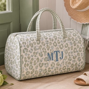 Könnte beinhalten: Eine gesteppte Reisetasche mit einem Leopardenmuster in Salbeigrün und Weiß. Die Tasche hat zwei Tragegriffe mit einem grün-weißen Vichy-Karomuster und ist mit den dunkelblau gestickten Monogrammbuchstaben MTJ auf der Vorderseite versehen.