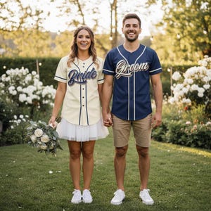 May include: A couple stands holding hands, wearing baseball jerseys. The jersey on the left is cream-colored with the word "Bride" in navy script. The jersey on the right is navy blue with "Groom" in white script. They are standing on green grass.