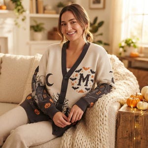 Könnte beinhalten: Eine cremefarbene Strickjacke mit Knopfleiste, dunkelgrauem V-Ausschnitt und Ärmeln. Das Strickmuster zeigt Halloween-Motive wie ein Spukhaus, einen Halbmond, Fledermäuse, orangefarbene Kürbisse und einen großen Buchstaben M mit Fledermäusen.