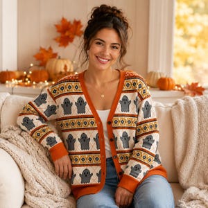 Könnte beinhalten: Eine cremefarbene V-Ausschnitt-Strickjacke mit einem sich wiederholenden Muster aus grauen Geistern sowie orangefarbenen und braunen geometrischen Bändern. Der geknöpfte Cardigan hat lange Ärmel mit gerippten orangefarbenen Bündchen und einen passenden Saum.