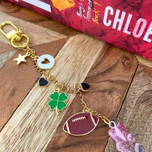 May include: Gold-tone keychain featuring charms: a star, a white heart-shaped disc, a black heart, a green shamrock, a burgundy American football, and a pink gummy bear. The keychain is presented on a wooden surface.