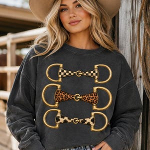 Könnte beinhalten: Dunkelgraues Sweatshirt mit drei goldfarbenen Pferdezahn-Designs. Die Gebisse haben karierte, Leoparden- und Kuhfellmuster. Das Sweatshirt wird mit einem beigefarbenen Cowboyhut und einer blauen Jeans getragen.