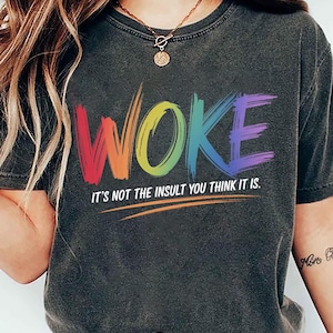 May include: A dark gray t-shirt with the word "WOKE" printed in rainbow colors. The text "It's not the insult you think it is." is printed below the word "WOKE".
