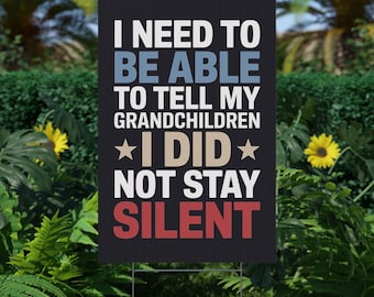 May include: A black yard sign with the text "I NEED TO BE ABLE TO TELL MY GRANDCHILDREN *I DID* NOT STAY SILENT" in white, blue, tan, and red. The sign is displayed outdoors in a garden setting.