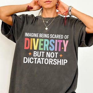 Op de afbeelding: Donkergrijs t-shirt met de tekst "IMAGINE BEING SCARED OF DIVERSITY BUT NOT DICTATORSHIP" in kleurrijke letters. Het shirt heeft een relaxte pasvorm en korte mouwen. De tekst is in een regenboogkleurenschema.