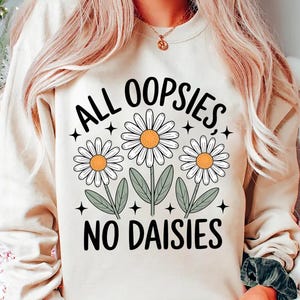 Puede incluir: Una sudadera color crema con el texto "ALL OOPSIES, NO DAISIES" en negro. El dise&ntilde;o presenta tres margaritas blancas con centros amarillos y hojas verdes. La sudadera es de estilo casual y c&oacute;modo.