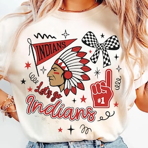 Könnte beinhalten: Cremefarbenes T-Shirt mit einem Grafikdesign, das einen indianischen Häuptling, einen Wimpel mit der Aufschrift "Indians", eine karierte Schleife und die Worte "Let's go Indians" zeigt. Das Design enthält auch rote Sterne und einen Schaumstoff-Finger.