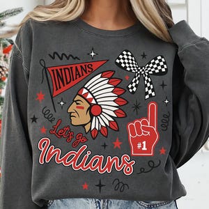 Könnte beinhalten: Dunkelgraues Sweatshirt mit einem grafischen Design. Es zeigt einen indianischen Kopfschmuck, ein Banner mit der Aufschrift "Indians", eine karierte Schleife und einen Schaumstofffinger. Der Text "Let's go Indians" ist ebenfalls enthalten.