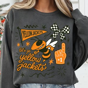 May include: Dark gray sweatshirt with a graphic design featuring a cartoon bee, a banner that says "YELLOW JACKETS", and the phrase "let's go yellow jackets". The design includes a checkered bow and a foam finger.