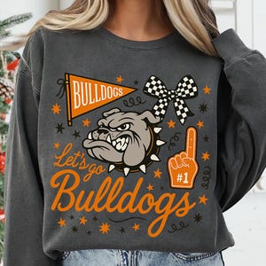 May include: Dark gray sweatshirt with a bulldog graphic and the words "Let's go Bulldogs." The design includes an orange pennant, stars, a checkered bow, and a foam finger. The sweatshirt is a comfortable, casual style.