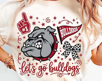 May include: Cream-coloured t-shirt featuring a bulldog graphic. The design includes a red #1 foam finger, stars, and a banner that says "BULLDOGS." The phrase "Let's go bulldogs" is written in a cursive font.